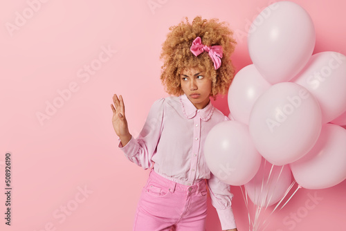 Unhappy young woman with curly hair wears blouse pink jeans and bowtie keeps palm raised and refuses something holds bunch of inflated balloons comes to congratulate best friend with birthday