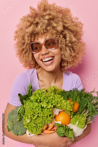 Vertical shot of positive woman with curly hair wears sunglasses embraces bunch of fresh green vegetabls poses with grocery against pink background. Healthy food and seasonal vitamins concept photo