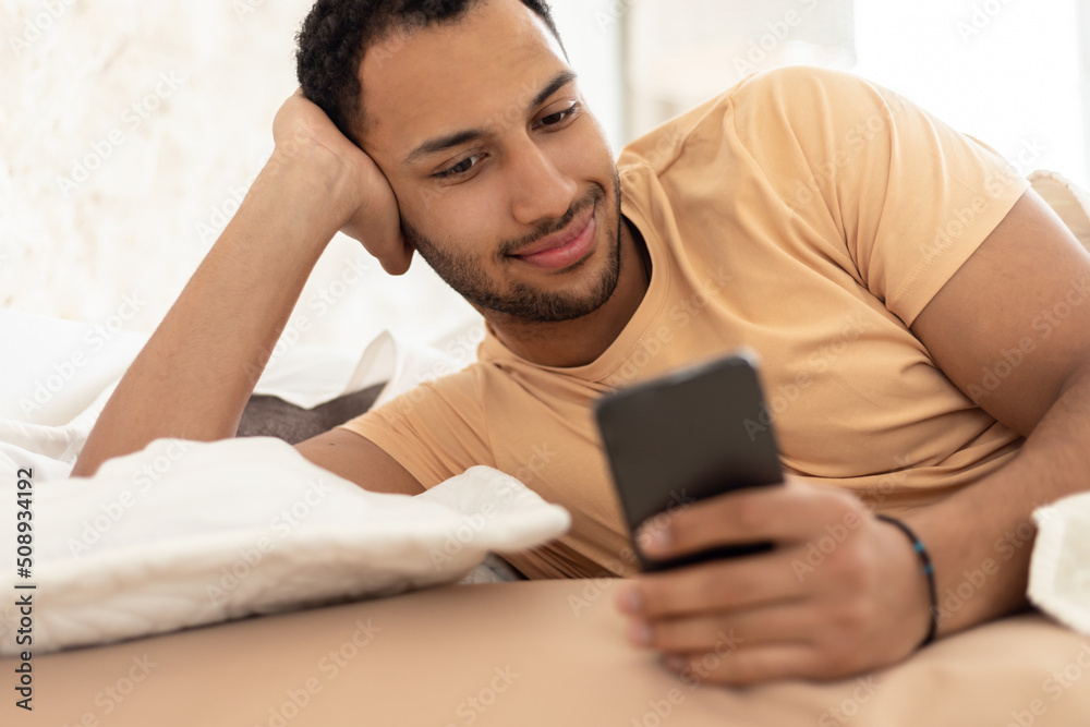 Middle Eastern Guy Using Mobile Phone Lying In Bed Indoor