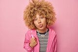Romantic lovely curly haired woman keeps lips folded wears casual striped jumper and jacket focused at camera wants to kiss you isolated over pink background. Human facial expressions concept