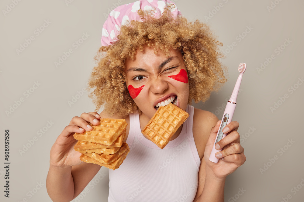 Photo of curly haired woman eats delicious homemade waffle holds electric brush for cleaning teeth applies red beauty patches to remove wrinkles dressed in casual t shirt isolated on grey studio wall - obrazy, fototapety, plakaty 
