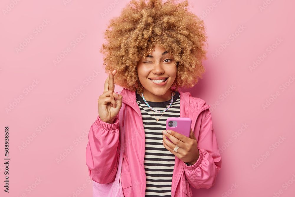 Beautiful hopeful curly haired woman holds mobile phone keeps fingers crossed believes in good luck winks eye dressed in casual jacket isolated over pink background. May my dreams come true.