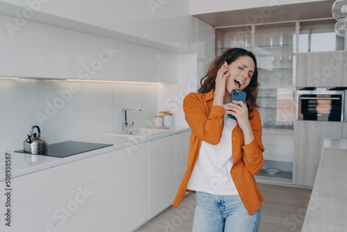 Happy housewife having fun with mobile telephone. Playful girl is dancing and singing alone at home.