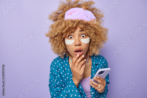 Stunned curly haired woman stares bugged eyes cannot believe in shocking news applies beauty patches under eyes dressed in pajama sleepmask holds mobile phone isolated over purple studio wall