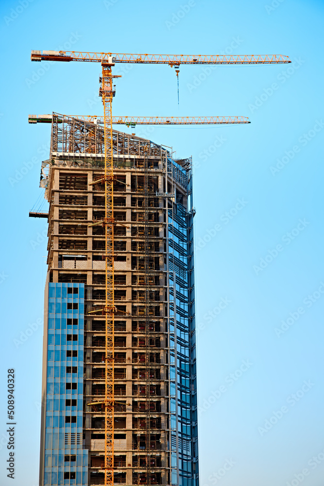 A cement and glass skyscraper under construction with two yellow tower cranes against a clear blue sky in sunny weather