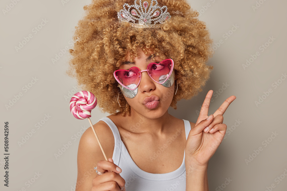 Dreamy lovely woman with curly blonde hair makes peace gesture with fingers holds lollipop applies silver patches under eyes dressed in t shirt isolated over grey background. Beauty concept.