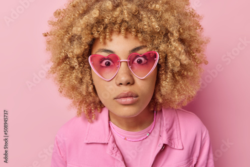 Close up portrait of shocked curly haired woman stares impressed at camera wears trendy sunglasses and jacket cannot believe own eyes isolated over pink background. Amazement and surprisement © wayhome.studio 
