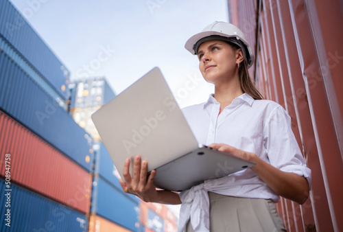 Dock manager or engineer worker in causual suit standing in shipping container yard holding laptop with smile. Import and export product. Manufacturing transportation and global business concept.
