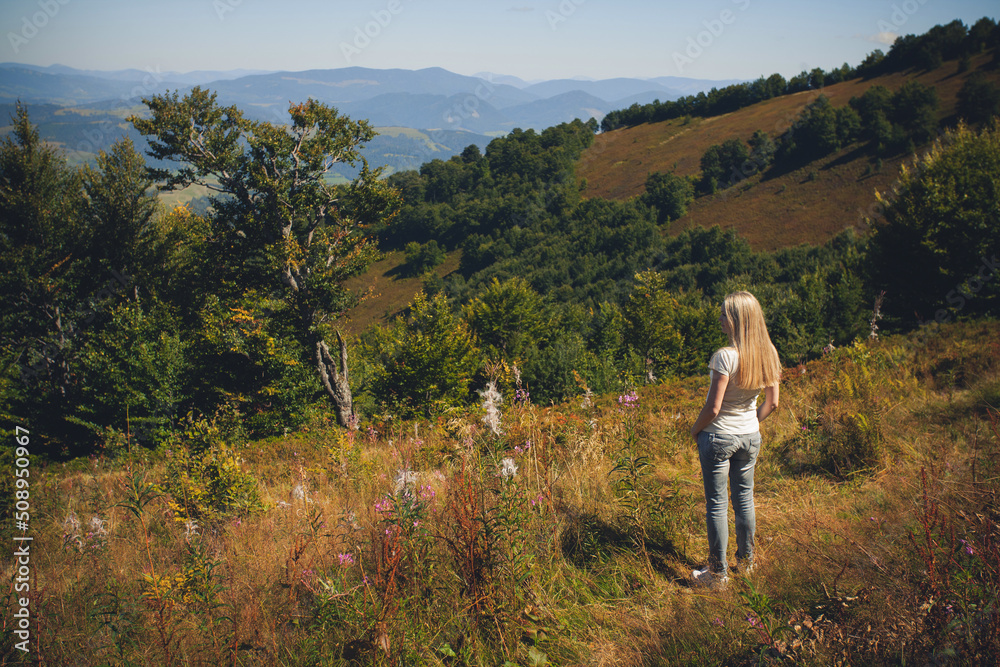 A slender young girl with long blond hair, in summer clothes, stands in the mountains, on a hot sunny day, and looks into the distance.