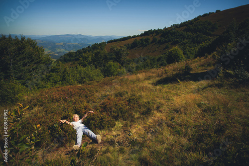 A little girl in blue pants and a light T-shirt lies in the grass in the mountains, in a hot summer. Around the grass, trees and blue sky with clouds.