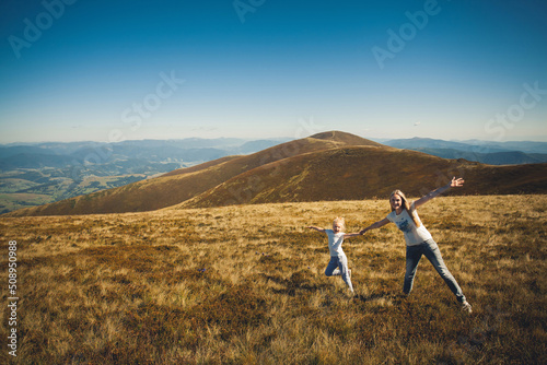 Happy family, mother and daughter, in summer clothes, having fun and laughing, holding hands and pulling each other on top of a mountain, on a sunny day in summer. photo