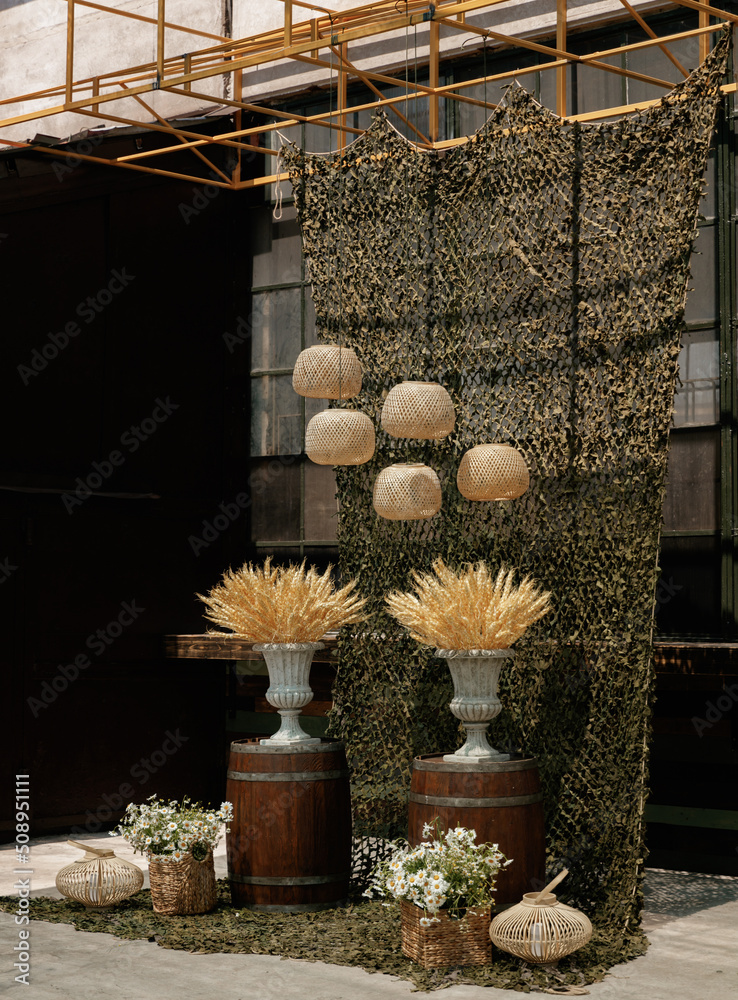 Obraz premium Wedding ceremony in a military style with wicker lamps, baskets with daisies, vases with dry wheat on wooden barrels