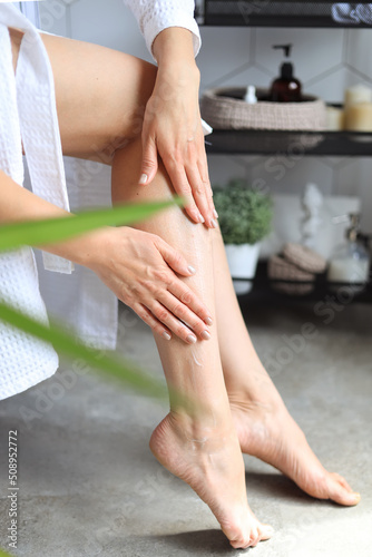 A young woman in a bathrobe touches the soft smooth skin of her legs, applying lotion, doing body care in the bathroom. Spa, laser epilation, depilation. Close-up