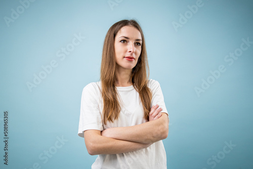 Thoughtful young woman in white t-shirt with arms crossed on blue background © Alex