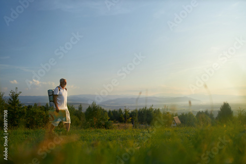 Strong middle aged man with backpack and map, enjoying sunset on top of hill outdoors. Side view of male tourist hiking alone, with picturesque mountain landscape on background. Concept of travelling.