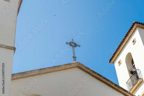 Detail of the metallic cross on the roof of a church, against blue sky with birds flying. 