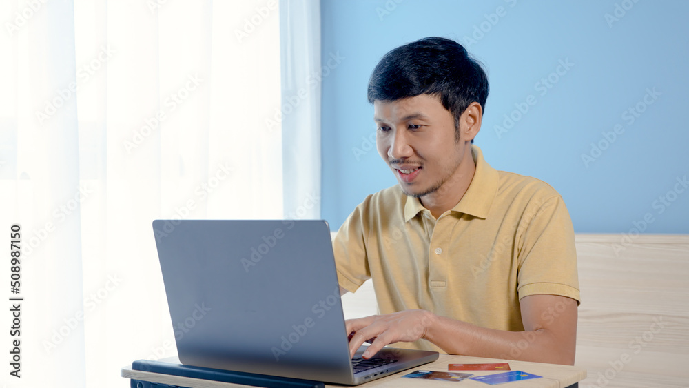 Handsome Asian man wearing a yellow shirt pays for online shopping on his laptop computer. The young man looked happy after completing his credit card payment. online shopping Handsome Asian man wearing a yellow shirt pays for online shopping on his laptop computer. The young man looked happy after completing his credit card payment. online shopping