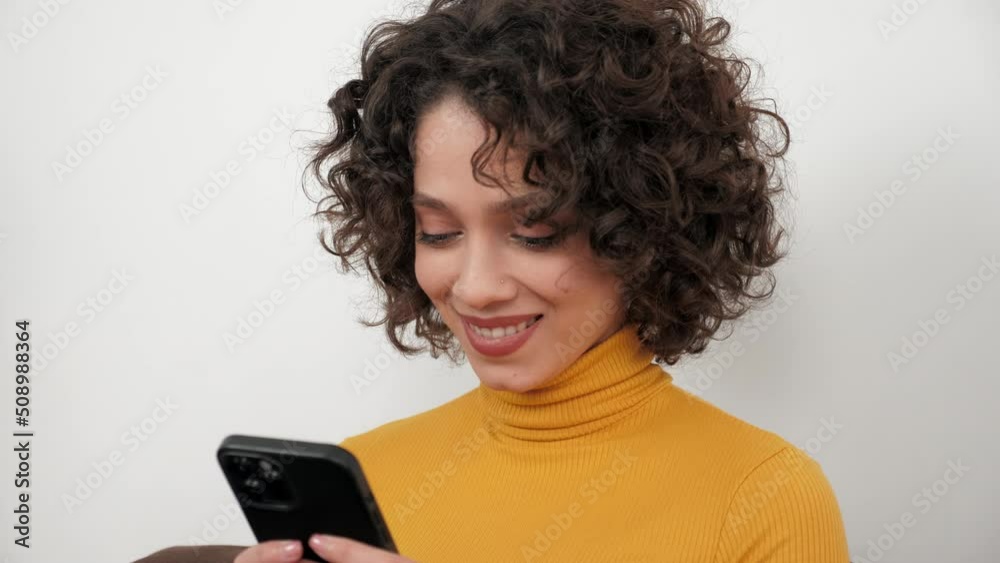 Smiling curly woman uses smartphone tapping screen display, sitting on couch at home. Positive businesswoman works for mobile phone online chatting with company employees, distance business meeting Smiling curly woman uses smartphone tapping screen display, sitting on couch at home. Positive businesswoman works for mobile phone online chatting with company employees, distance business meeting