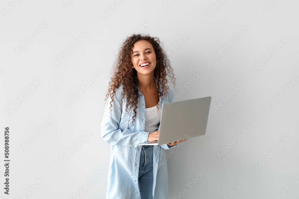Happy young African-American woman using laptop on light background - obrazy, fototapety, plakaty 