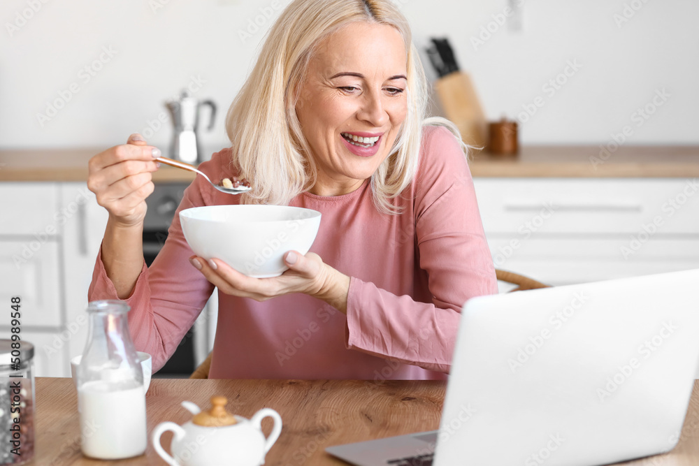 Morning of mature woman with laptop having breakfast in kitchen Morning of mature woman with laptop having breakfast in kitchen