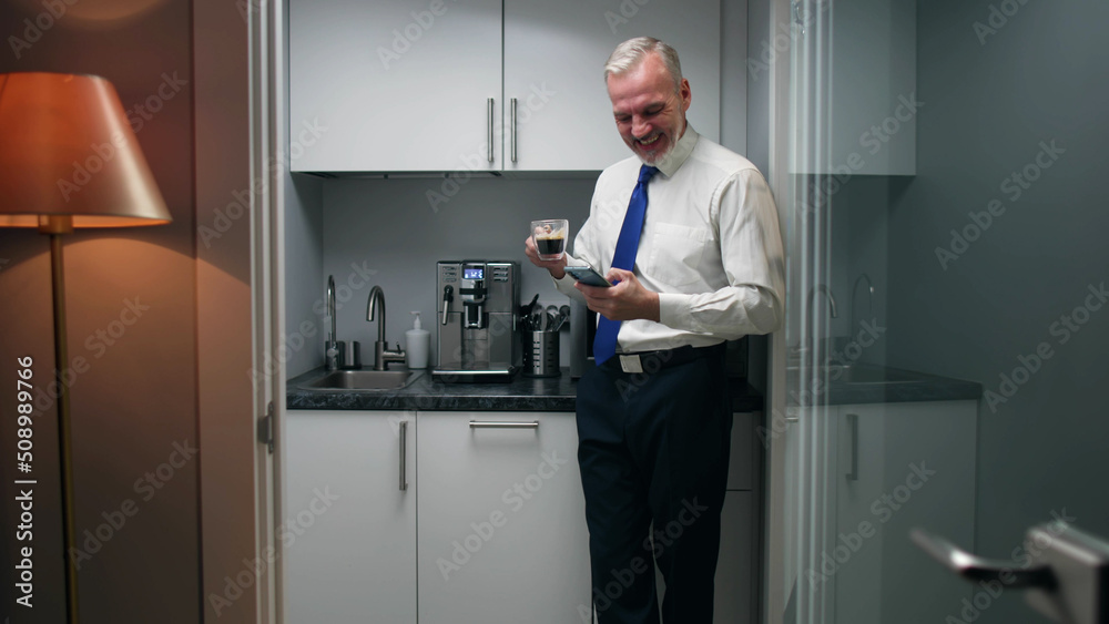 Senior executive drink coffee and use smartphone standing in corporate kitchen