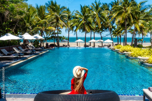 Luxury beach vacation in tropical beach hotel. Tourist woman in red dress relax near blue swimming pool in modern resort. Female traveler on sea vacation.