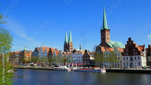 malerische Häuser entlang der Trave in Lübeck mit Kirchtürmen und Ausflugsbooten bei blauem Himmel