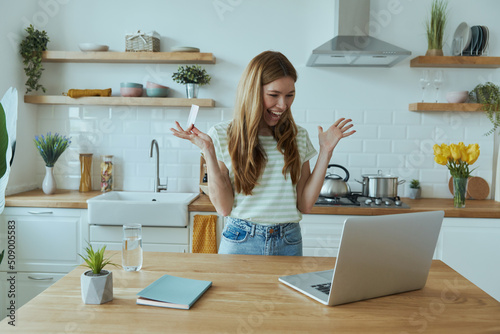 Excited young woman enjoying online shopping and using credit card while sitting at the kitchen