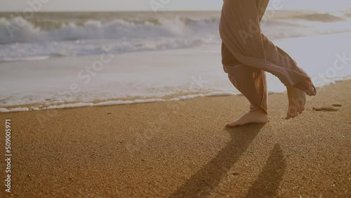 A young woman walks by the sea at sunset. Legs of a girl against the background of waves and the sea. Sportswear. Barefoot on the sand