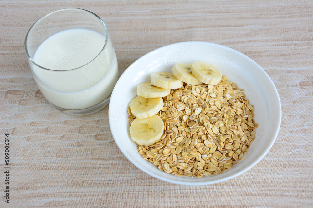 oatmeal with banana slices in white bowl with glass of milk on beige background oatmeal with banana slices in white bowl with glass of milk on beige background