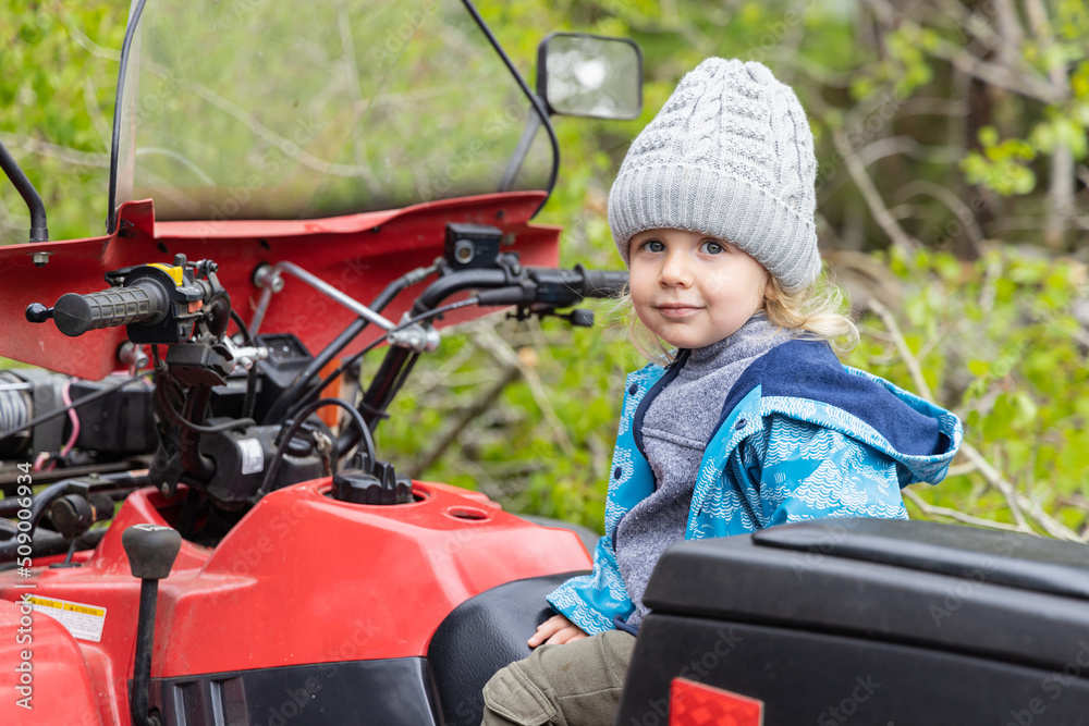 A happy three year old boy is seen in the driver seat of a red all terrain vehicle with handlebars in the countryside. With blurry foliage background.