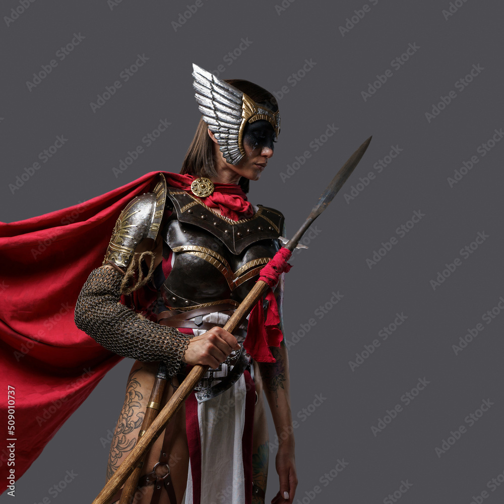 Obraz premium Shot of female barbarian holding spear dressed in steel armor with helmet against grey background.