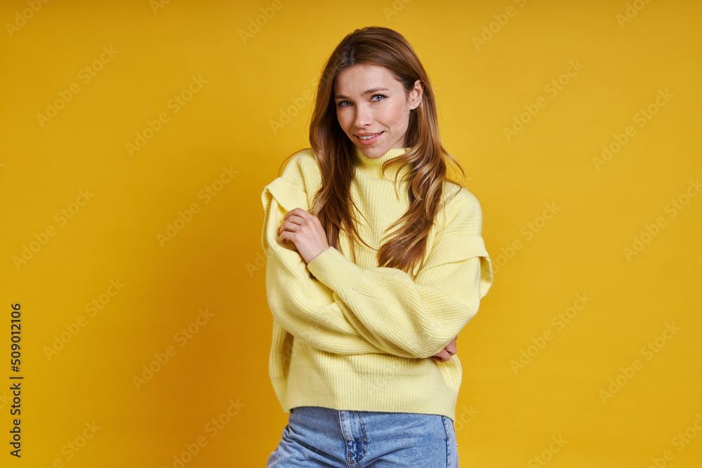 Beautiful young woman in cozy sweater hugging self while standing against yellow background Beautiful young woman in cozy sweater hugging self while standing against yellow background