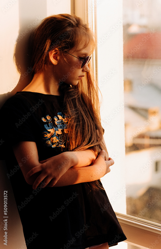 A little girl sits stands on the windowsill in a black T-shirt and sunglasses with her long hair loose at a golden sunset