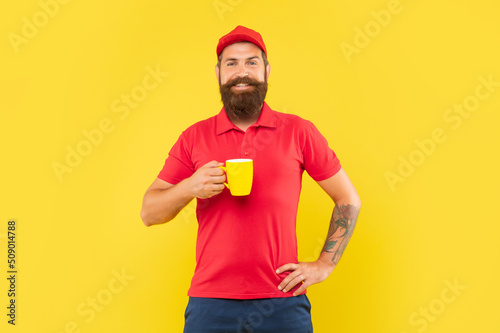 Happy bearded man in casual red cap and tshirt holding mug yellow background, coffee