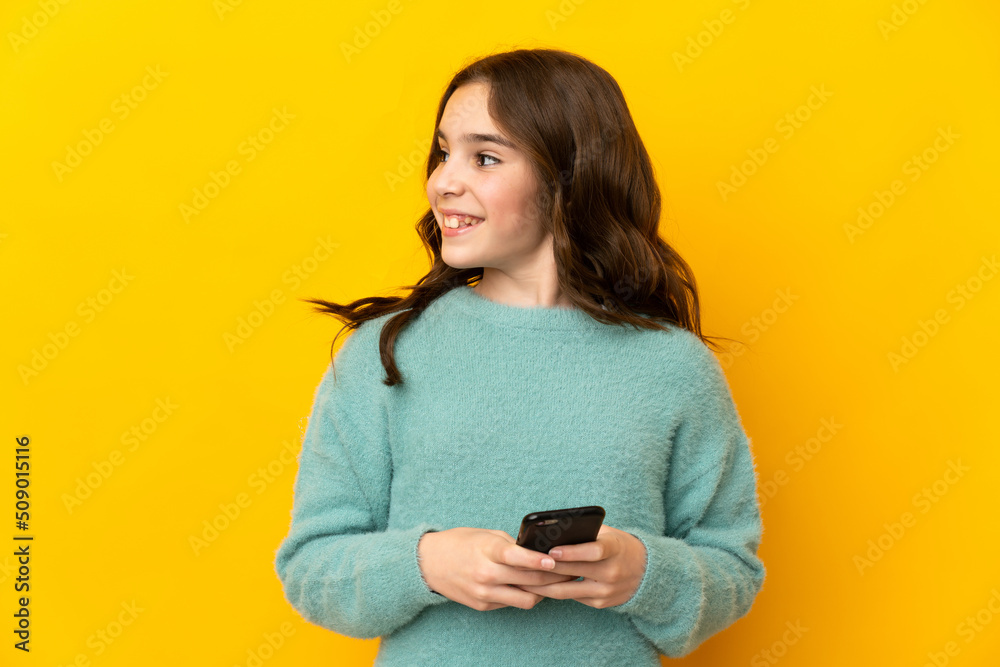Little caucasian girl isolated on yellow background using mobile phone and looking up Little caucasian girl isolated on yellow background using mobile phone and looking up