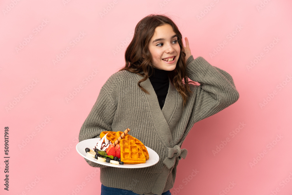 Little girl holding waffles isolated on pink background listening to something by putting hand on the ear Little girl holding waffles isolated on pink background listening to something by putting hand on the ear