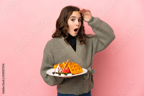 Little girl holding waffles isolated on pink background doing surprise gesture while looking to the side