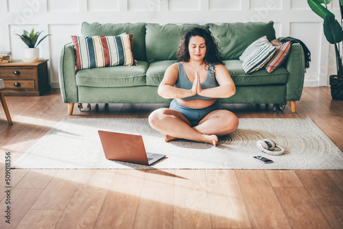 Curly haired overweight young woman in top and shorts turns on online yoga training and practices exercises on floor mat against green sofa by wall