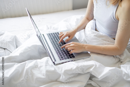Happy casual beautiful woman working on a laptop sitting on the bed at the morning in the house. Working at home  freelance concept.