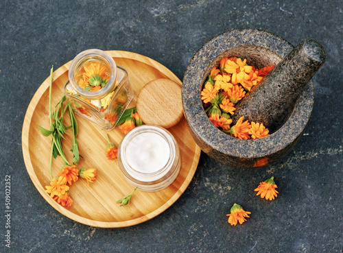 Calendula Cream for body care in glass jar and fresh orange calendula flowers on a wooden plate and a granite mortar on black table. Concept: spa, aromatherapy, Medical Dermatology. Flat lay