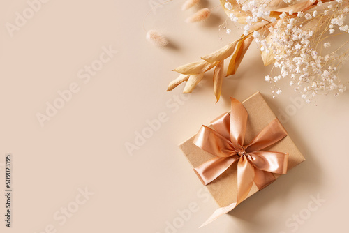 Gift box with golden ribbon and dry grass and flowers on beige background
