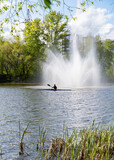 A girl on a kayak boat floats past a river fountain in the city, which splashes water high up.
