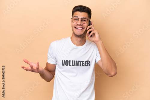 Young volunteer caucasian man isolated on beige background keeping a conversation with the mobile phone with someone