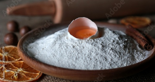 Rustic style still life of organic food ingredients for homemade baking with white flour and egg yolk inside half of broken egg shell. Flour powder pile in traditional wooden bowl. Dried orange slices