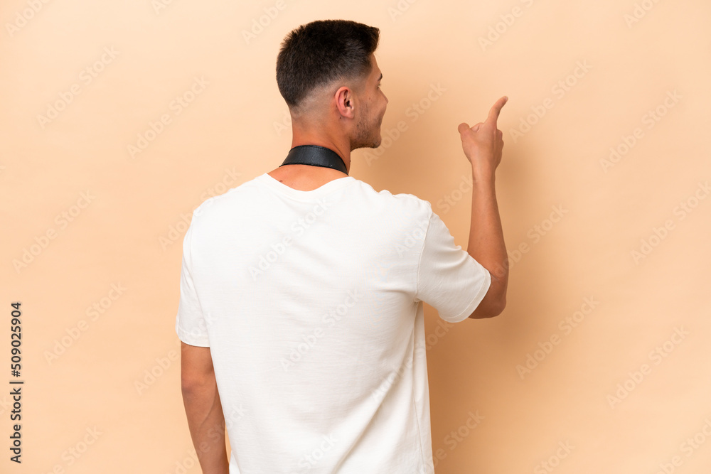 Young photographer caucasian man isolated on beige background pointing back with the index finger