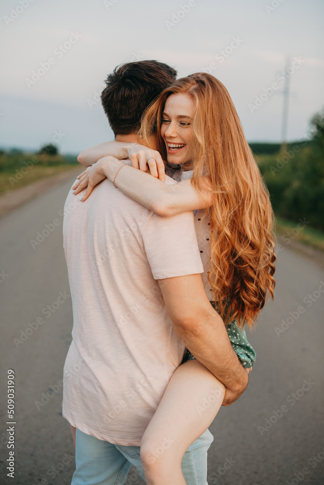 Man carrying his ginger girlfriend on shoulder while walking by the country road, the girl laughs. I will steal you. St. Valentine's Day. Lovers holiday