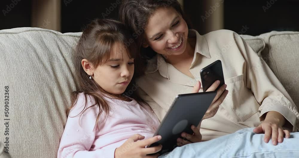 Family use diverse gadgets at home, close up. Mother, cute little daughter sit on couch with digital tablet and smart phone spend time on internet, modern wireless tech overuse, new apps usage concept Family use diverse gadgets at home, close up. Mother, cute little daughter sit on couch with digital tablet and smart phone spend time on internet, modern wireless tech overuse, new apps usage concept