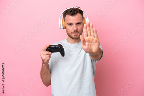 Young Brazilian man playing with video game controller isolated on pink background making stop gesture