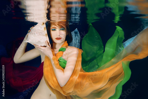 Redhead woman with big sea shell in colorful clothes on the dark background underwater